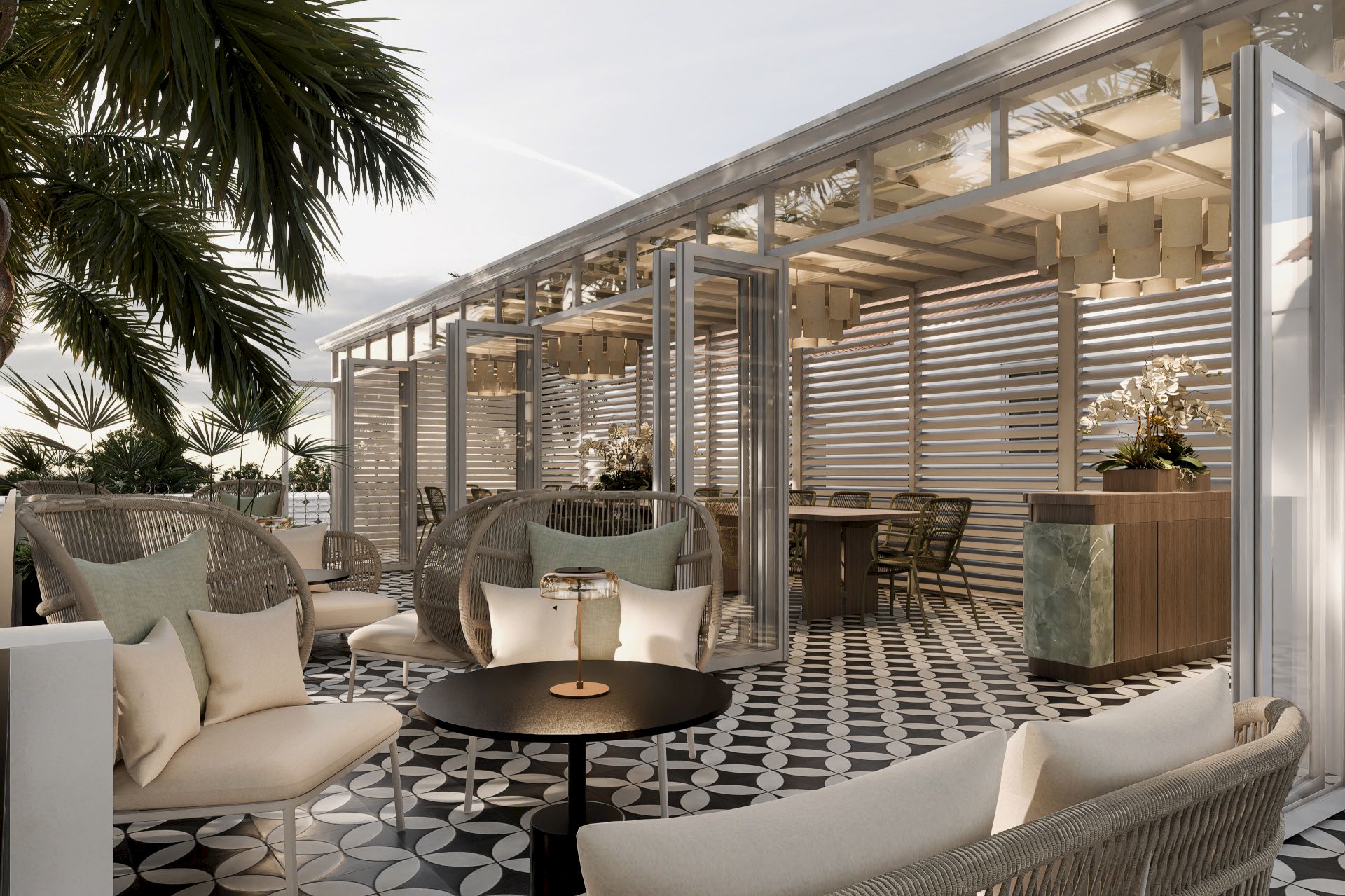 A modern outdoor lounge with beige sofas, a round coffee table, patterned tiles, a glass pergola, and palm trees in view.